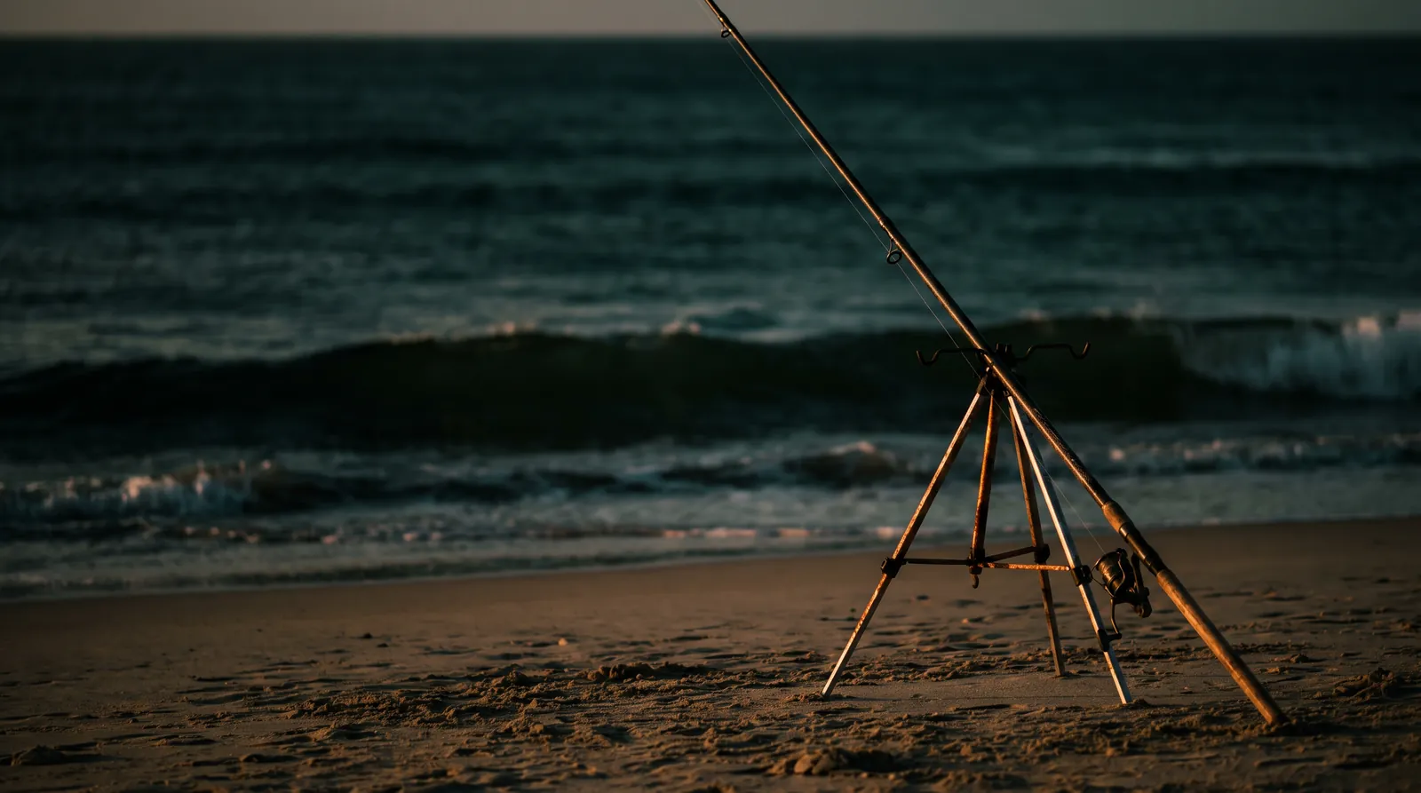 Une longue canne de surfcasting dressée dans un trépied sur une plage de sable à l'aube, des vagues basses derrière