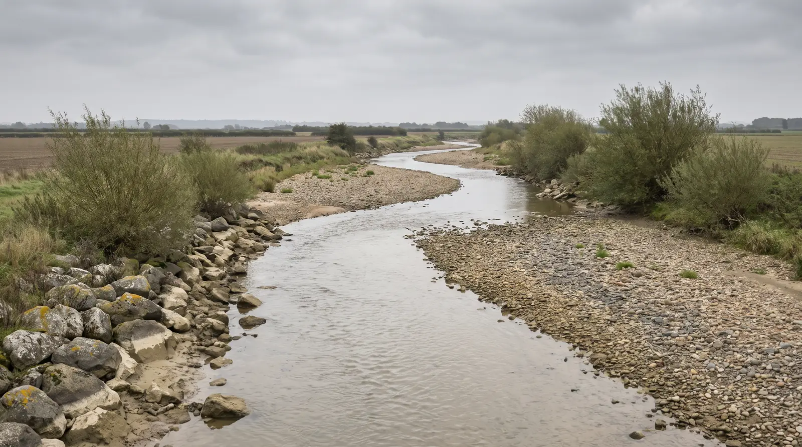 Une rivière d'étiage sévère réduite à un ruban d'eau étroit mais continu, serpentant entre de larges bancs de galets à découvert, une bande claire marquant l'ancien niveau au bas des blocs.