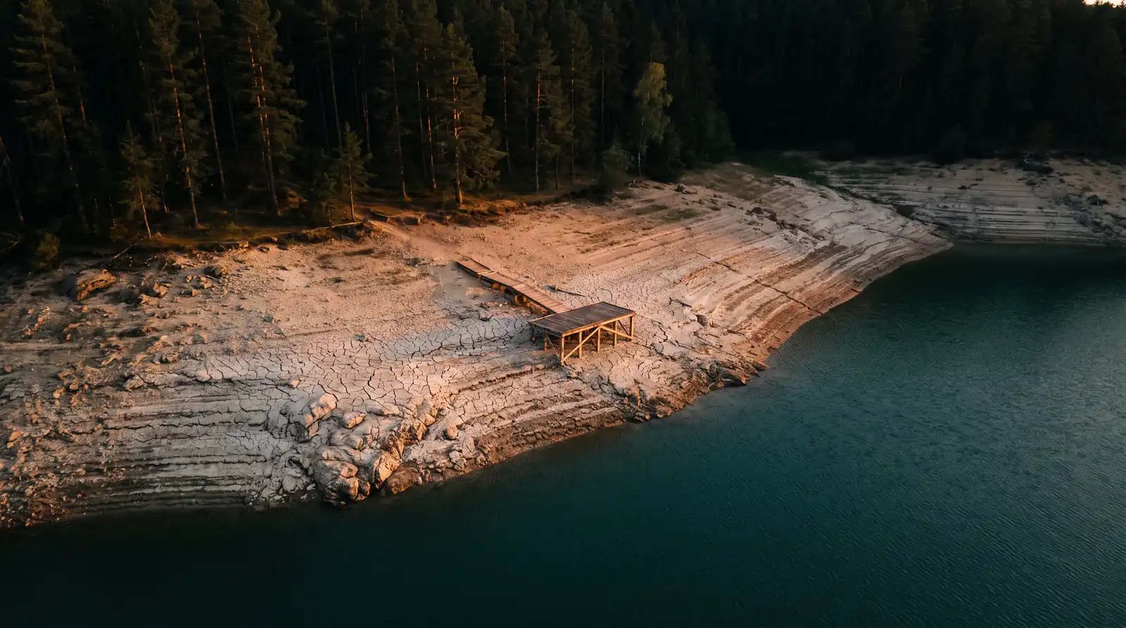Un lac de barrage très bas au crépuscule, une large bande de rive sèche et craquelée entre la forêt et l'eau, un ponton resté à sec